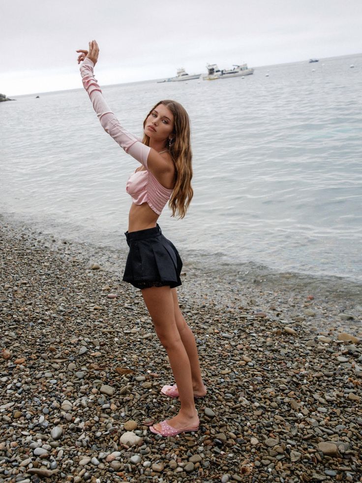 a young woman, possibly a teenager or young adult, stands on a pebbly beach with her arms raised. she is wearing a light pink, longsleeved crop top and black shorts. she's also wearing pink sandals.  the ocean is in the background, with some boats visible in the distance. the sky is overcast and the overall mood is calm and relaxed.  the image appears to be a fashion photograph.