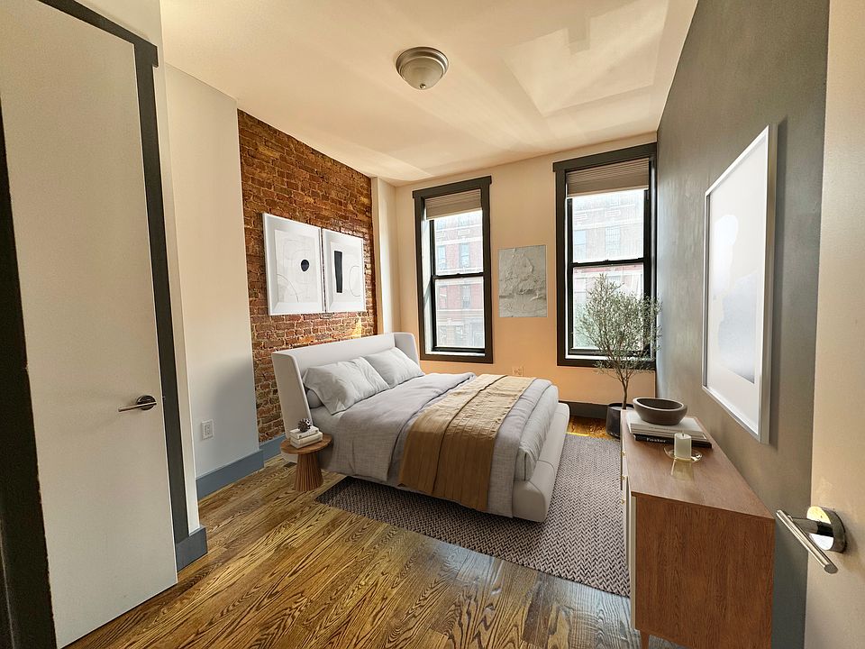 this is a nicely furnished bedroom with a bed, bedside table, and a wooden chest of drawers. the room has wooden floors, a brick wall, and two windows with black frames. there is a small potted plant next to the chest of drawers, a wall art hanging above the chest of drawers and a couple of wall arts hanging above the bed. the room also has a ceiling light.