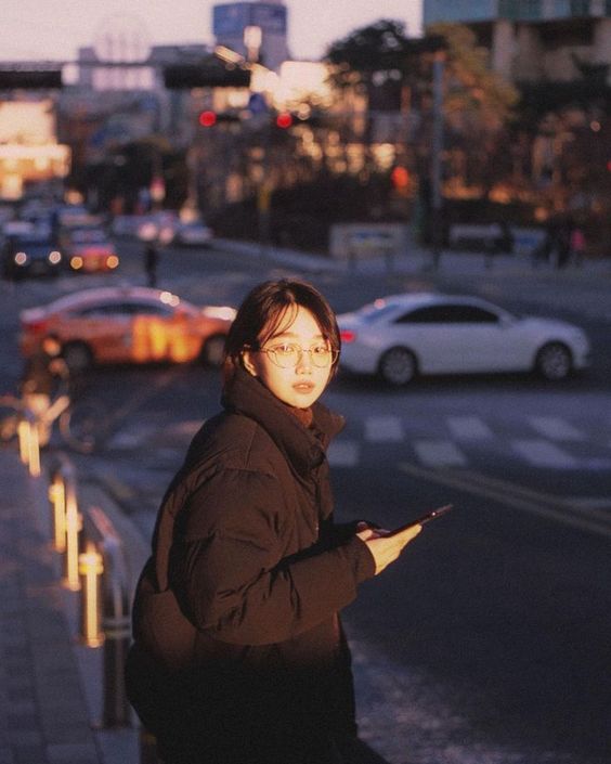 a young woman stands on a city street corner, bathed in the warm glow of the setting sun. she is wearing a black puffer jacket and round eyeglasses, and she is holding a smartphone in her hand. cars drive by in the background, and the city lights are starting to twinkle. the image has a warm, nostalgic feel.