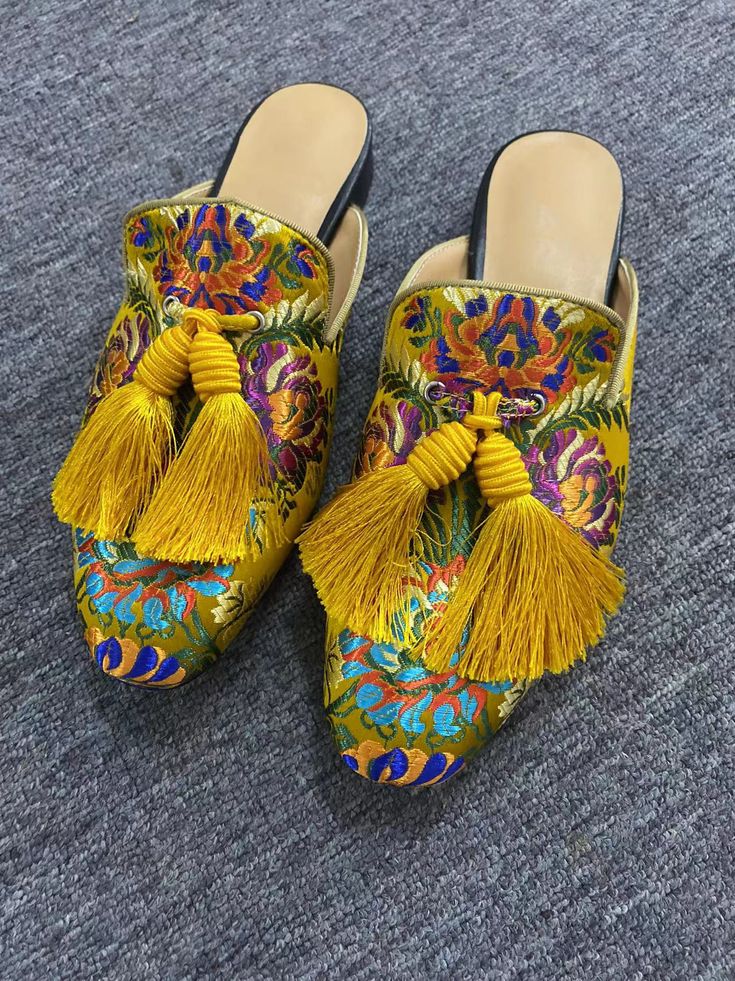 a pair of women's embroidered mules with yellow tassels are shown on a dark gray carpet. the mules are made of a yellow brocade fabric with colorful floral embroidery. the tassels are a bright mustard yellow.  the shoes appear to be handmade or artisanmade. the image is taken from a slightly above angle, showing both shoes clearly.