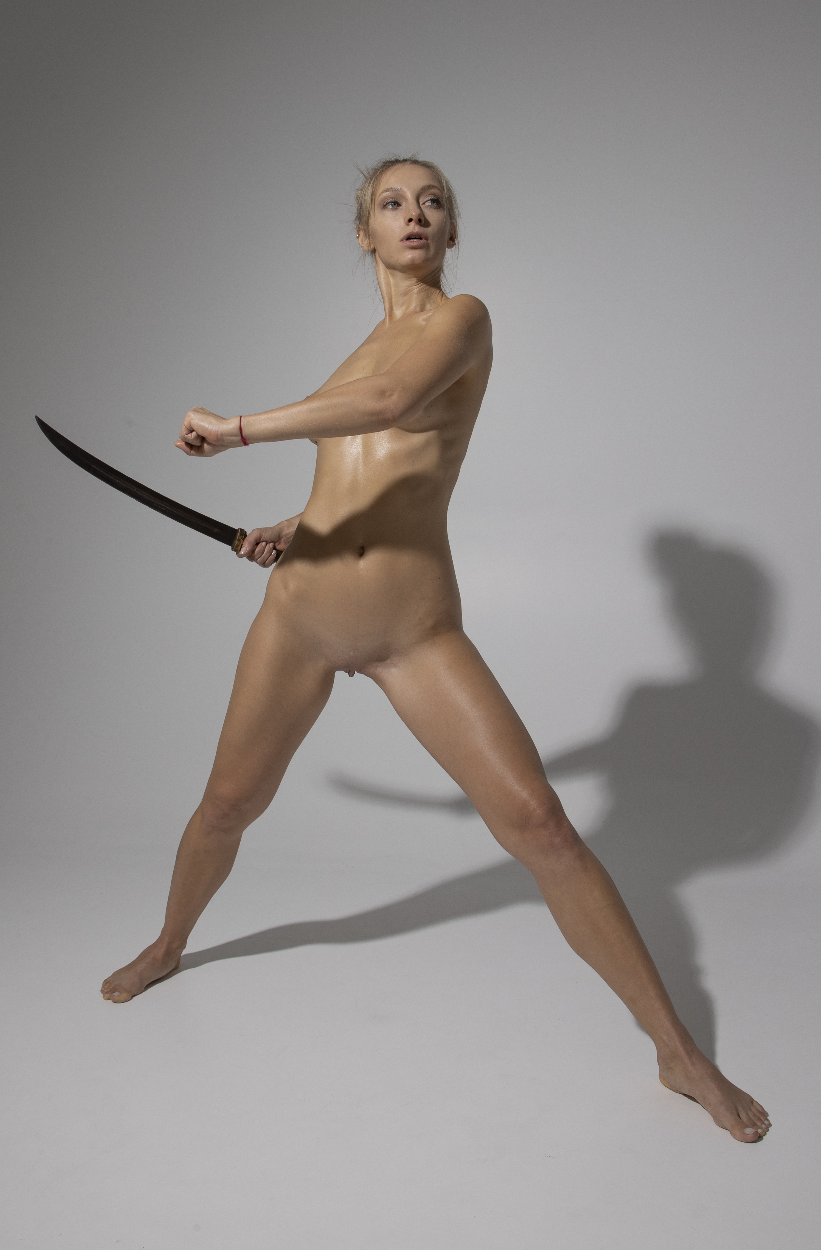 a nude woman is standing in an open stance with a sword in her left hand. she is lightskinned and appears to have oiled skin. a shadow is cast behind her. she is looking to the right.