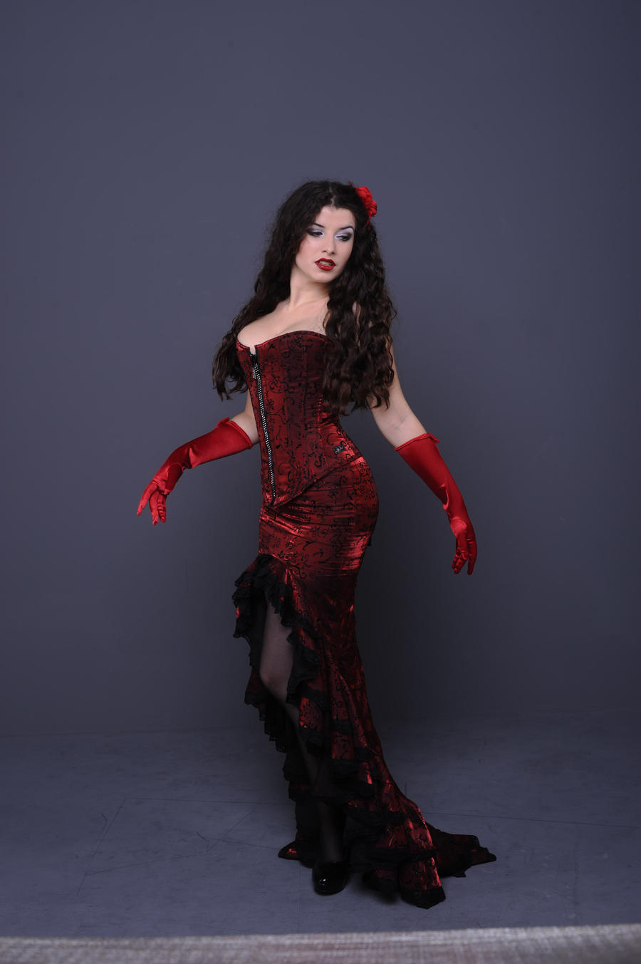 a fulllength studio shot of a woman with long dark curly hair. she is wearing a dramatic, long red velvet dress with a corset top and black lace detailing. the dress features a high slit revealing black stockings and high heels. she's wearing long red gloves and a small red flower is tucked into her hair.  the background is a muted gray. the overall style is elegant, possibly vintage or burlesque inspired, with a sensual and confident pose.
