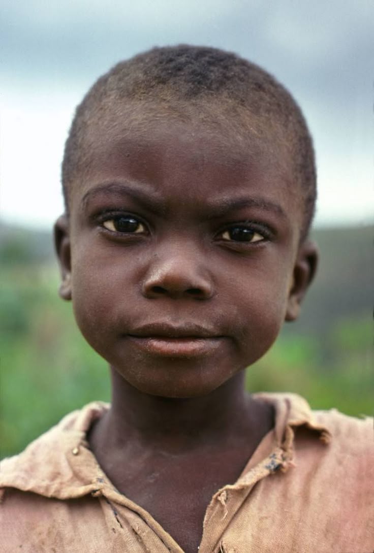 a closeup portrait of a young african boy with dark skin, short hair, and a serious expression on his face. he is wearing a brown shirt. the background is blurred and green.