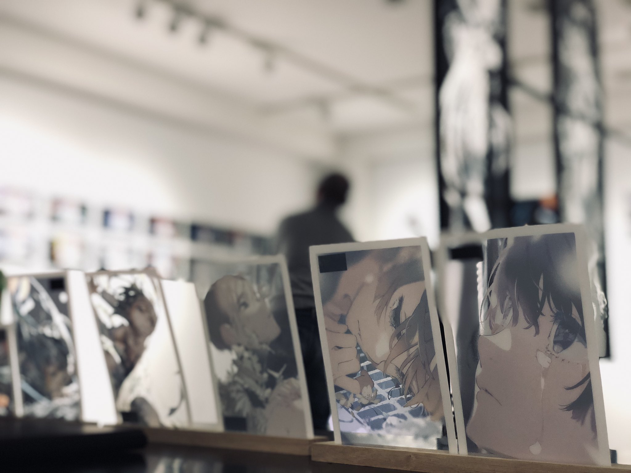 the image showcases a closeup view of several mangastyle illustrations displayed on a wooden stand within an art gallery. the artwork features characters with expressive eyes and detailed linework.  the gallery setting is suggested by the white walls, track lighting, and a blurred figure of a person in the background, likely an artist or visitor admiring the exhibition. the overall ambiance is bright and inviting, drawing attention to the intricate details of the illustrations.