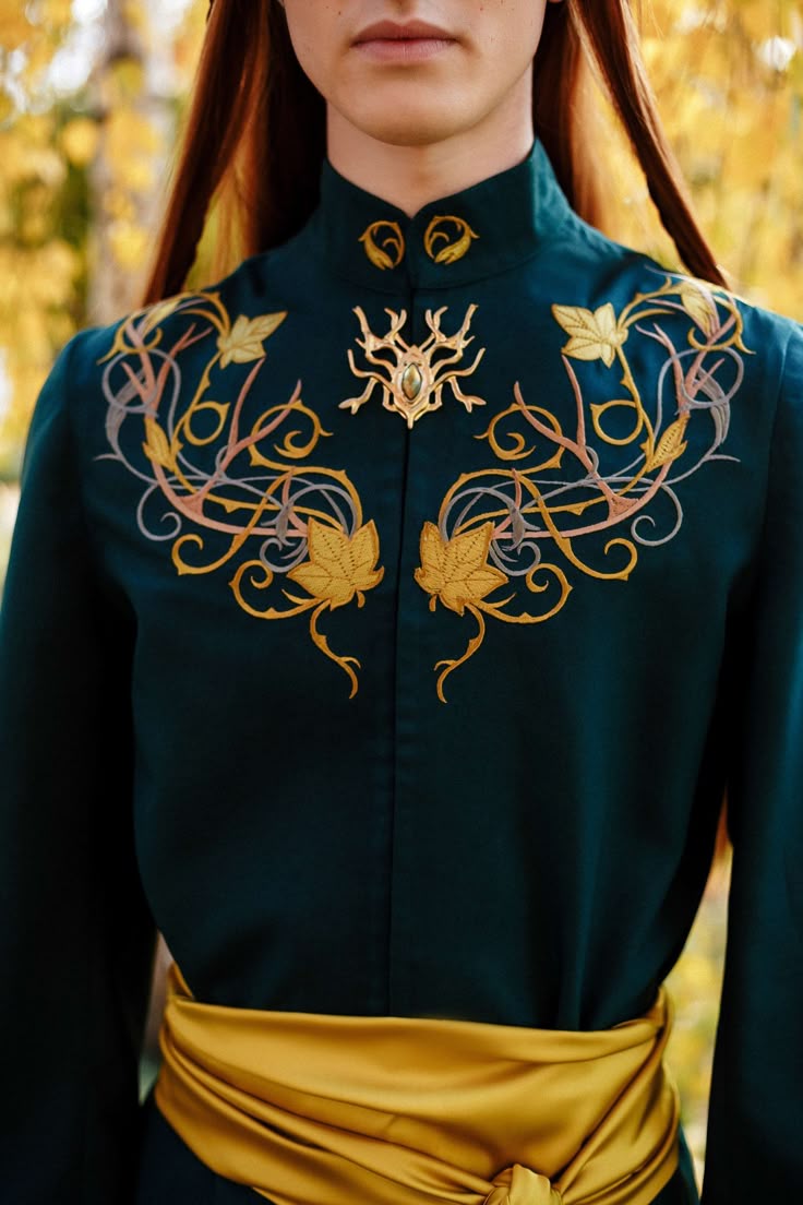 a person in an elven costume is depicted in a closeup shot. the costume features a dark green tunic with intricate gold embroidery of leaves and vines on the upper chest. a golden sash is tied around the waist. a decorative brooch is pinned at the center of the chest, just below the neck. the person has fair skin and long redbrown hair. the background is blurred, suggesting an outdoor setting with trees.