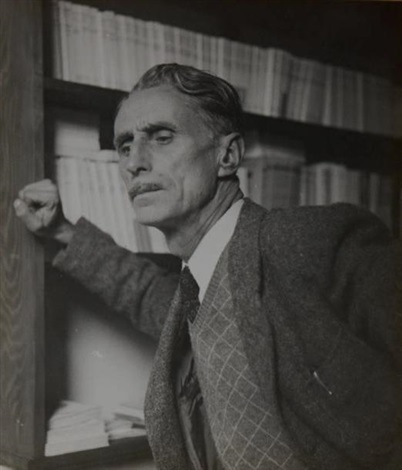 the image is a black and white photograph of an older man, possibly in his 50s or 60s. he appears to be of european descent, and his expression is serious and contemplative. he is dressed in a tweed jacket and a collared shirt with a tie, suggesting a somewhat formal or professional setting. the man is leaning against a large wooden bookshelf filled with numerous books, suggesting a studious or academic environment. the background is somewhat out of focus, but it is clearly a wellstocked library or study. the lighting is soft and the overall mood of the photograph is quite pensive and intellectual.  the photo's style and quality suggest it might be a vintage print from the mid20th century.