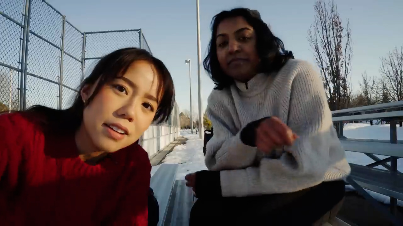 two young women are taking a selfie while sitting on a park bench in the winter. the sun is setting behind them, casting a warm glow on the snowcovered ground. one woman is wearing a red sweater and the other is wearing a white sweater. they are both smiling and appear to be enjoying each other's company.