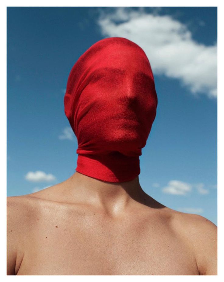 a woman with red fabric covering her head and face. the fabric is seethrough, so you can see her features but they are blurred. the woman is wearing a red fabric around her neck. the background is a blue sky with white clouds. the woman is looking straight ahead.