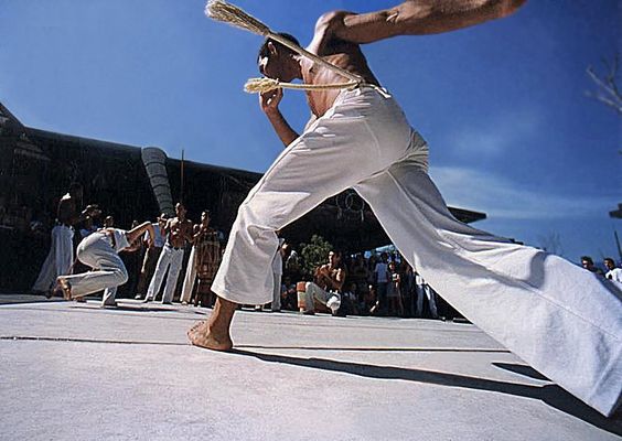 two men performing capoeira in an outdoor space. the man in the foreground is midair, about to kick the other man. a large crowd is watching. the photo is taken from a low angle. the day is sunny with a clear blue sky.