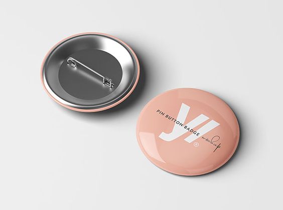 the image shows two round pin button badges, one facing up and the other facing down. they appear to be made of metal and have a glossy finish. the badge facing up is pink and features the text "pin button badge" and "mockup" in a light script font. the other badge shows the back of the button and its pin. both badges are set against a light gray background.