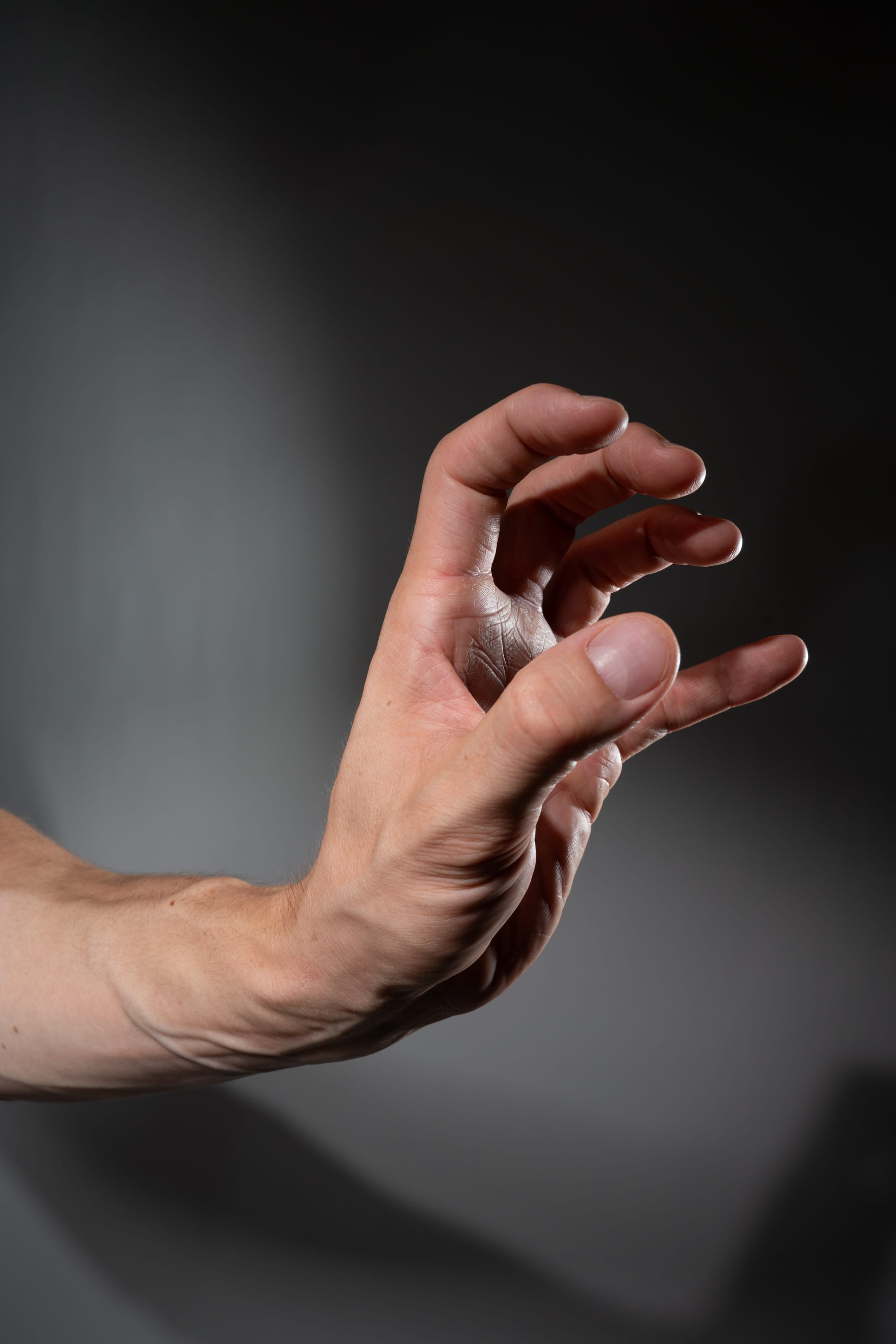 a man's right hand is reaching forward with fingers slightly curled as if grasping for something.  the image is a closeup shot of the hand and arm against a dark gray background. the hand is lit with studio lighting highlighting the details and texture of the skin.