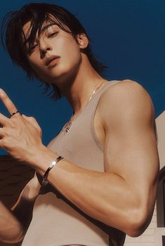 a closeup shot of a handsome young man with dark hair and brown eyes. he has a stylish, androgynous look and is wearing a simple tank top and silver jewelry, including a bracelet and a ring. the background is a soft blue. the lighting is artificial and soft, creating a flattering portrait.