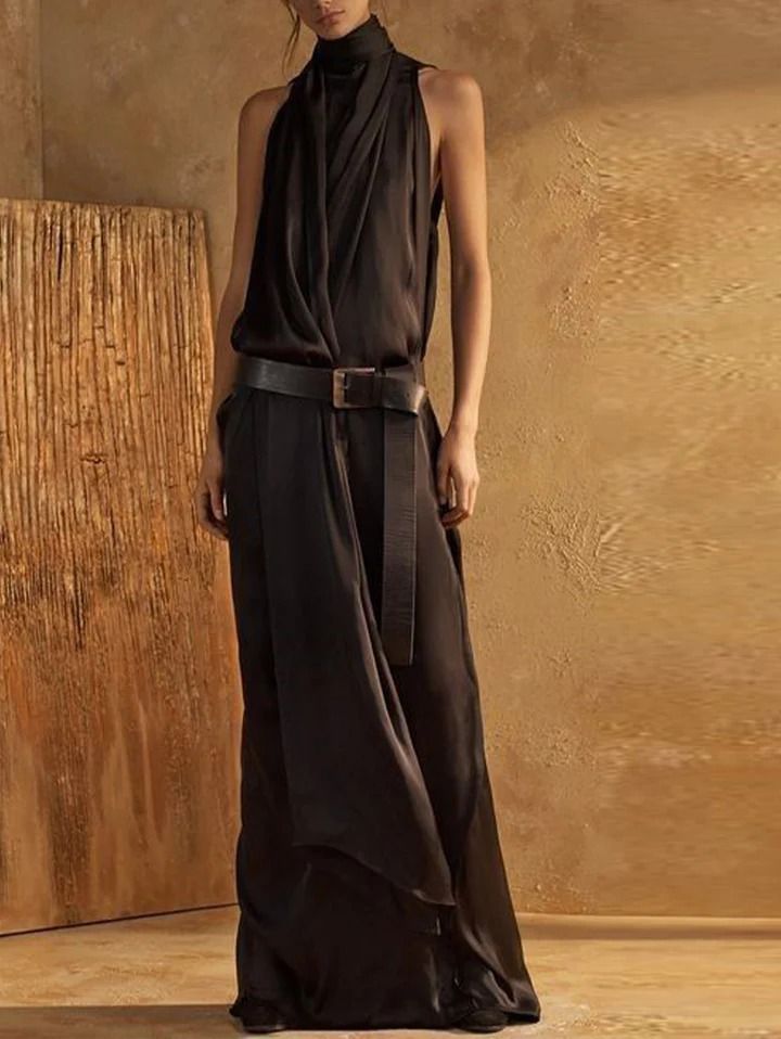 a woman stands against a brown wall wearing a long black sleeveless dress. the dress has a high neck, a belt, and a flowy design. she is posing with her hands on her sides. the background is a brown wall.