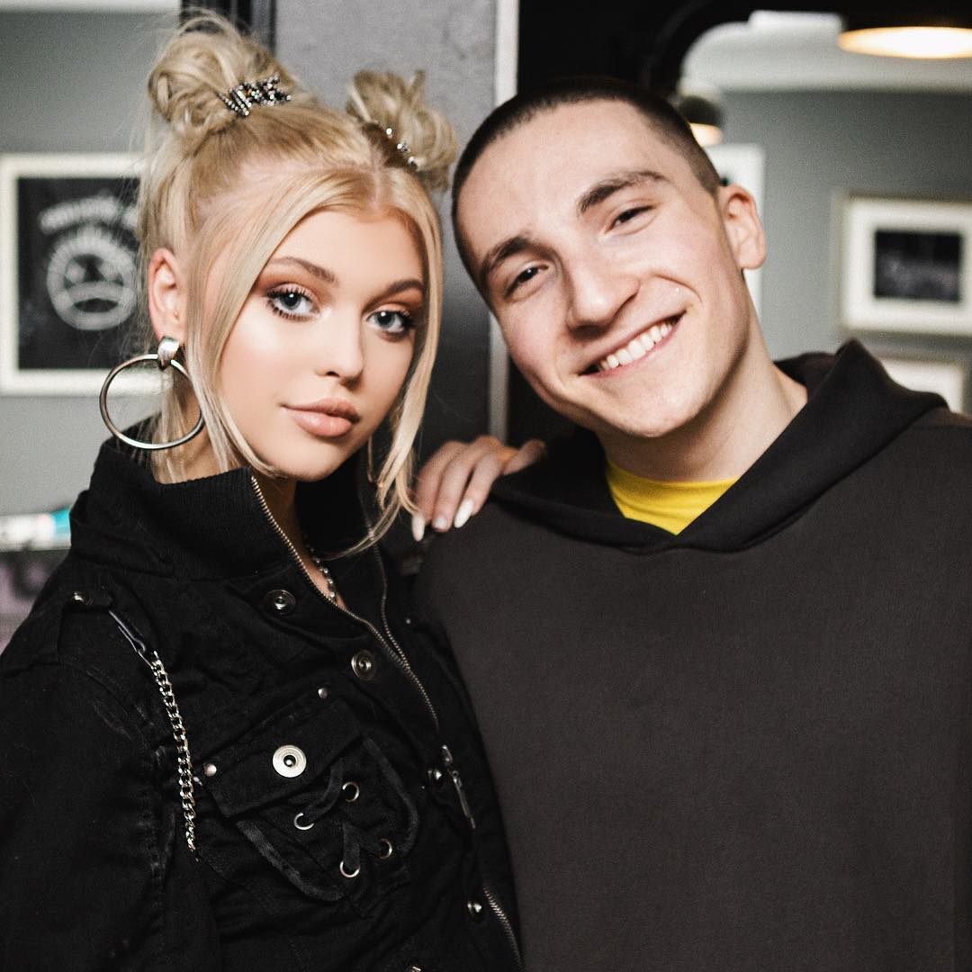 a photo featuring a blonde woman with her hair styled in space buns, wearing a black jacket and large hoop earrings, standing beside a smiling man in a gray hoodie with a yellow tshirt underneath.