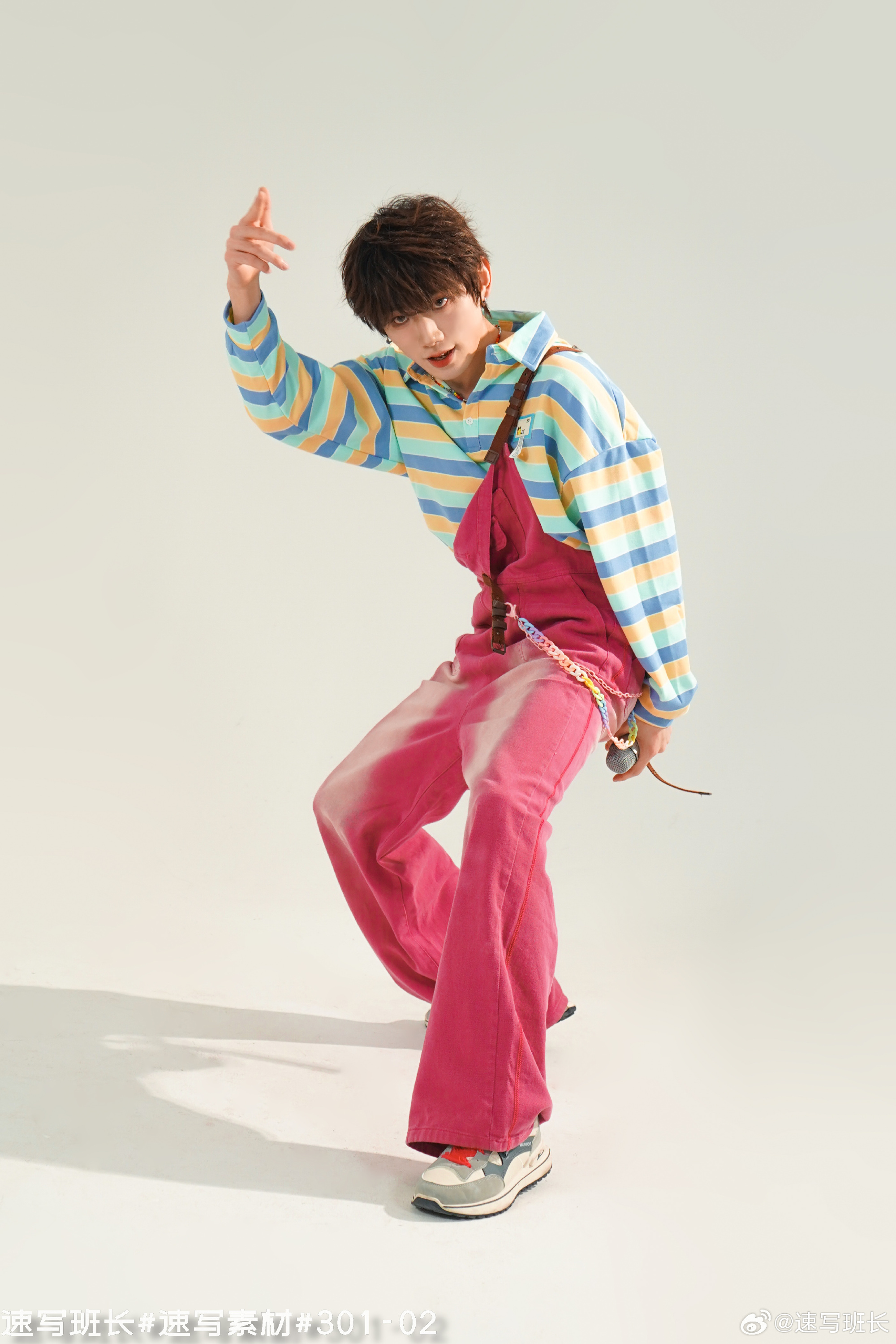 a young asian boy wearing colorful clothes is posing against a white background.  he is wearing a pastel striped sweater with blue collar and cuffs, pink overalls and gray sneakers. the boy is in midmotion, striking a pose that suggests he might be dancing or singing.