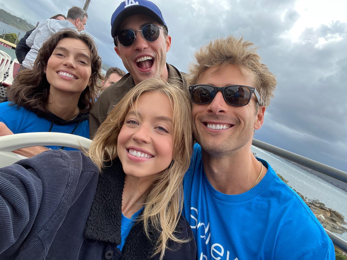 the image is a selfie taken outdoors on what appears to be a tourist bus.  four young adults are smiling happily for the photo. two women and two men are visible. the background shows a cloudy sky and a view of a body of water with a cityscape in the distance. the two women are positioned close to the camera, while the men are behind them.  all four are wearing casual clothing; one man is wearing a los angeles dodgers baseball cap, and two of the individuals are wearing sunglasses. the atmosphere is jovial and suggests friendship and enjoyment of a travel experience.