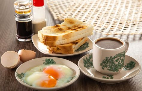 the image shows a classic malaysian or singaporean breakfast spread.  the main components are kaya toast (slices of toasted bread with kaya, a coconut jam, spread between them), two softboiled eggs, and a cup of kopi (traditional coffee).  condiments such as soy sauce and pepper are also visible.  the food is served on vintagestyle patterned plates and saucers. the overall setting appears homely and inviting.