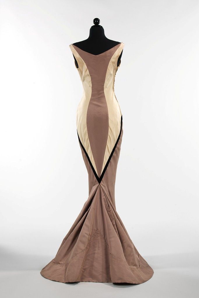 a gown is displayed on a black mannequin against a white background. the gown features a combination of brown and beige panels arranged in a geometric color block design. the dress is sleeveless and floorlength with a mermaid silhouette, flaring out near the bottom. black trim accentuates the geometric design.