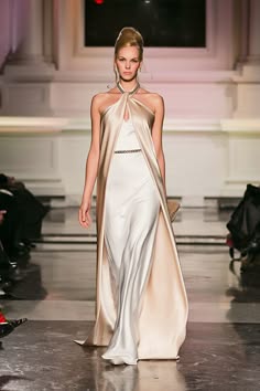 a female model wearing a white gown with a cream overlay is standing on a runway, posing for the camera. the gown is elegant and flowing, showcasing the model's beauty. the model is facing the camera and has a confident expression on her face. she is wearing minimal makeup and her hair is styled in a simple updo. the background is blurry, allowing the model and her dress to take center stage. this image is a classic example of fashion photography, showcasing the beauty of a stylish gown and the elegance of the model.