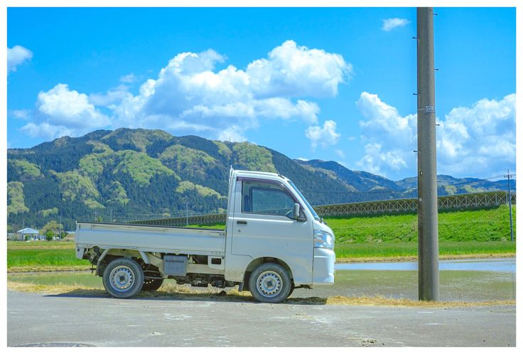 a white mini truck is parked on a paved road in a rural setting. in the background are mountains, a blue sky with white clouds, green grass, a paddy field, and a utility pole.