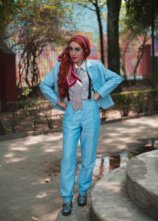 a full shot of a woman posing outdoors in a light blue pantsuit. she has long red hair tied back with a red headband and is wearing a red and white patterned tie, suspenders, and dark shoes.  she is standing on a stone walkway with steps in the foreground, and trees and greenery are visible in the background.  the woman appears confident and stylish.  the overall aesthetic is vintage or retro, possibly reminiscent of the 1970s or 1980s.