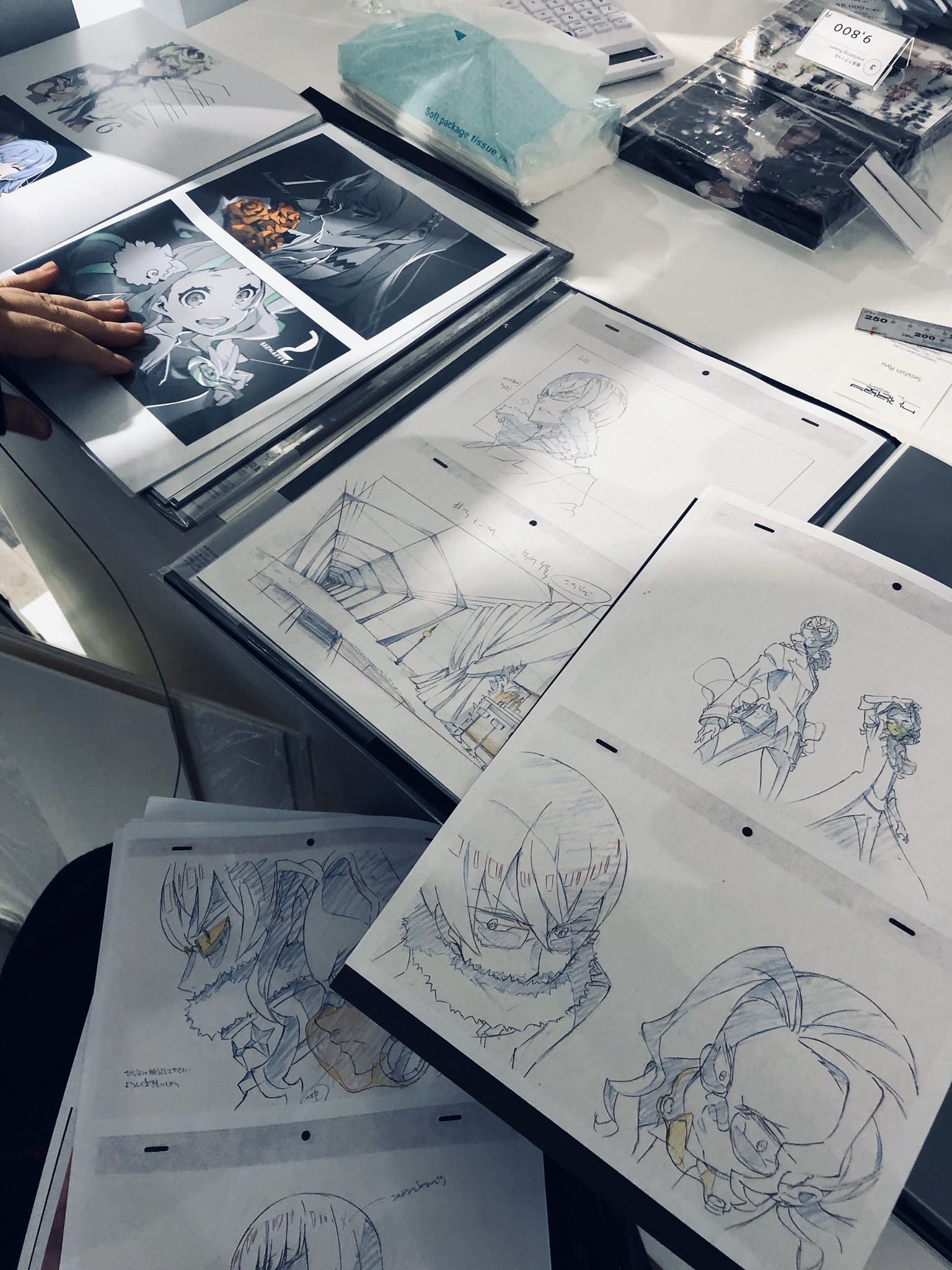 the image shows a table covered in art materials, likely belonging to an anime artist. open sketchbooks and loose drawings are scattered across the surface, displaying character designs and scene compositions in various stages of completion. the hand of an individual is visible flipping through the pages of a book showcasing more refined character illustrations. the scene is bathed in soft, natural light, highlighting the intricate details of the pencil work.