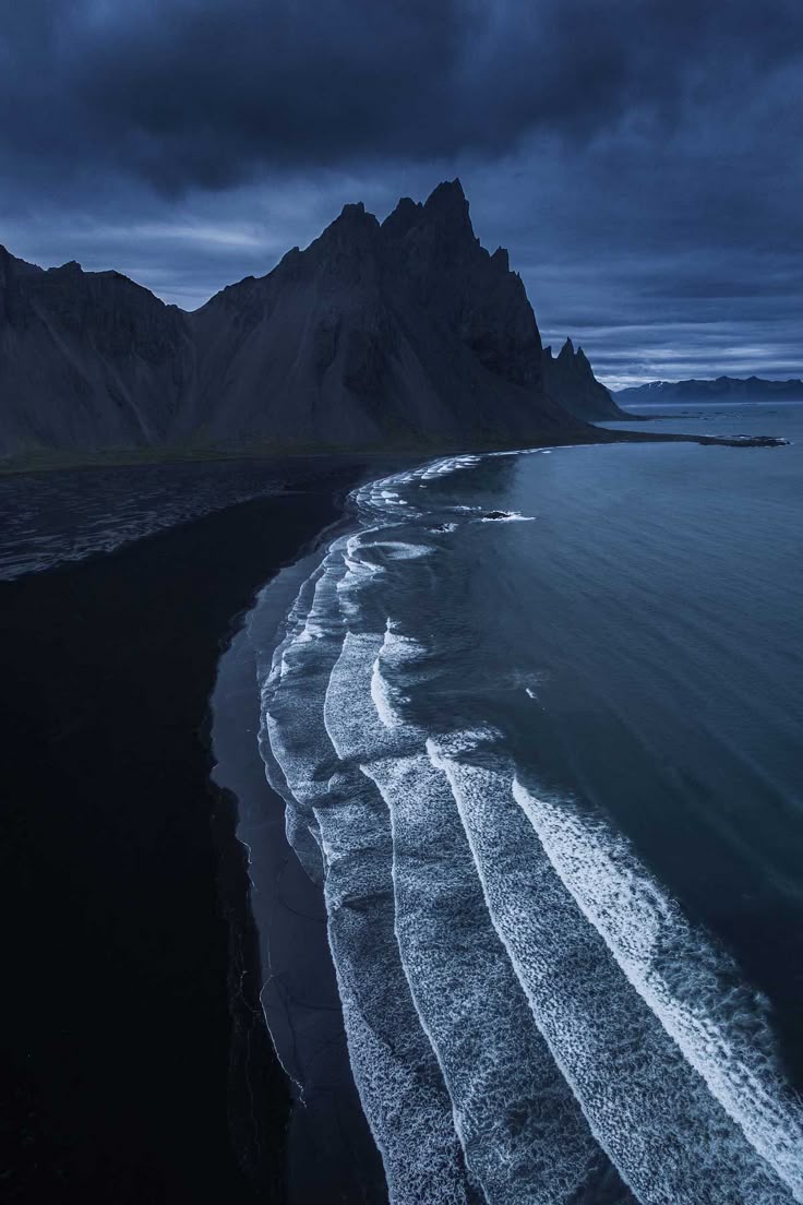 an aerial view of a dark landscape with a black sand beach, ocean, mountain, and cloudy sky. the waves are crashing on the beach. the image appears to be taken in stokksnes, iceland.