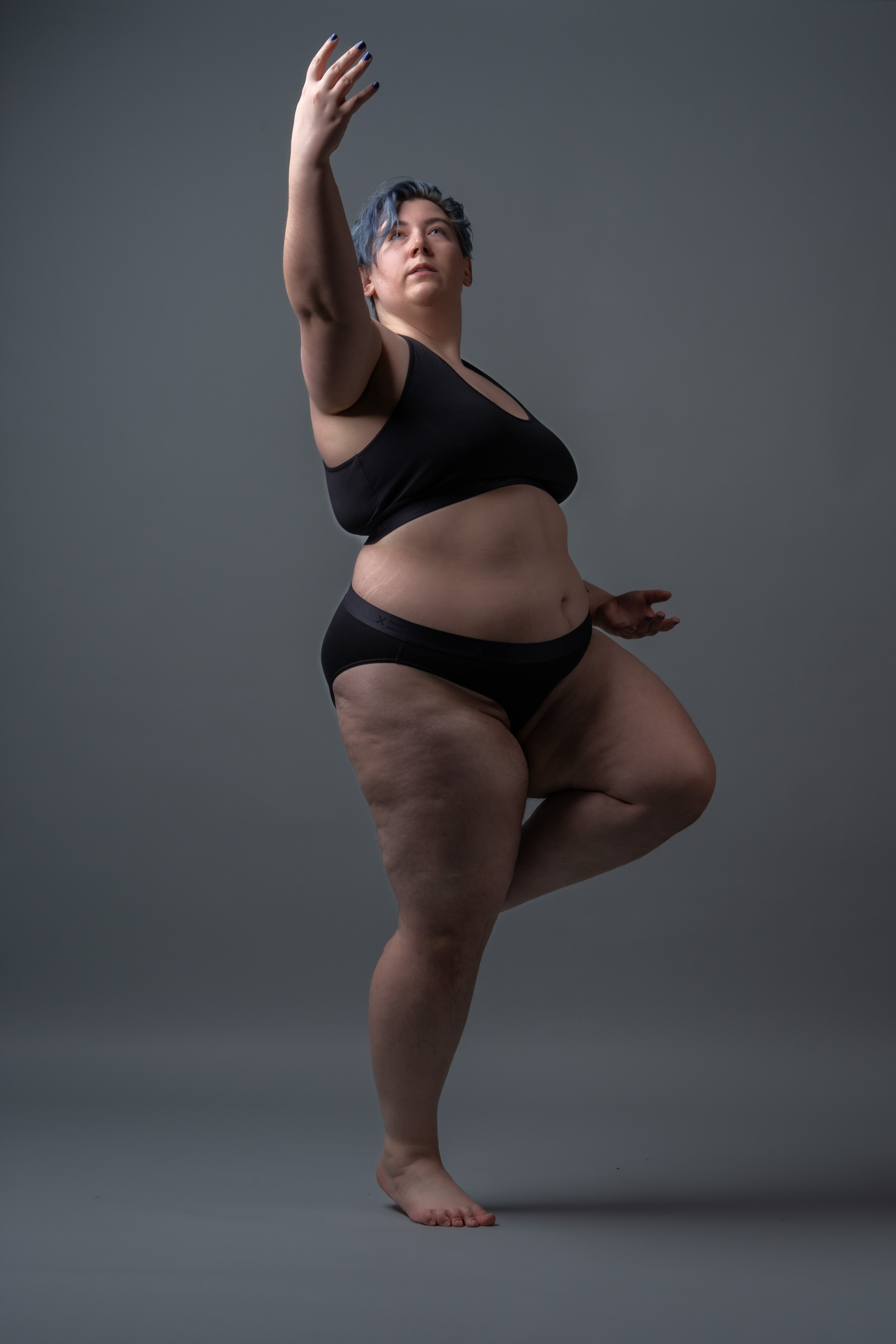 a plus size woman in black underwear poses in a studio against a gray background. she is standing on one leg with the other lifted, arm raised above her head and short blue hair.