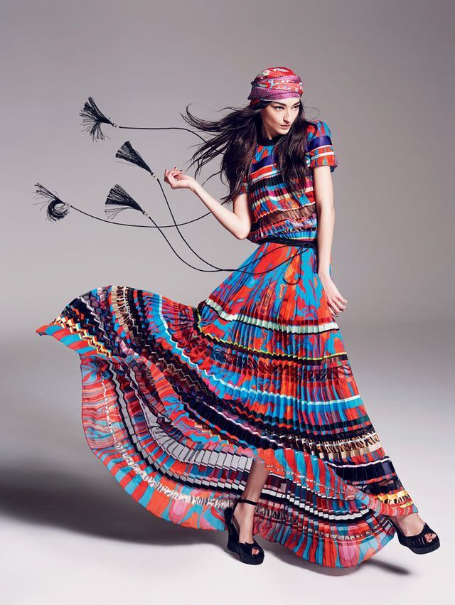 a fashion model is posing in a colorful long dress and sandals. the dress is pleated and flowy and the model's hair is long and flowing. she is wearing a black head scarf and has a bright red and blue striped pattern on her dress. the model is posing in a studio setting with a white background. the image is taken from a slightly low angle.
