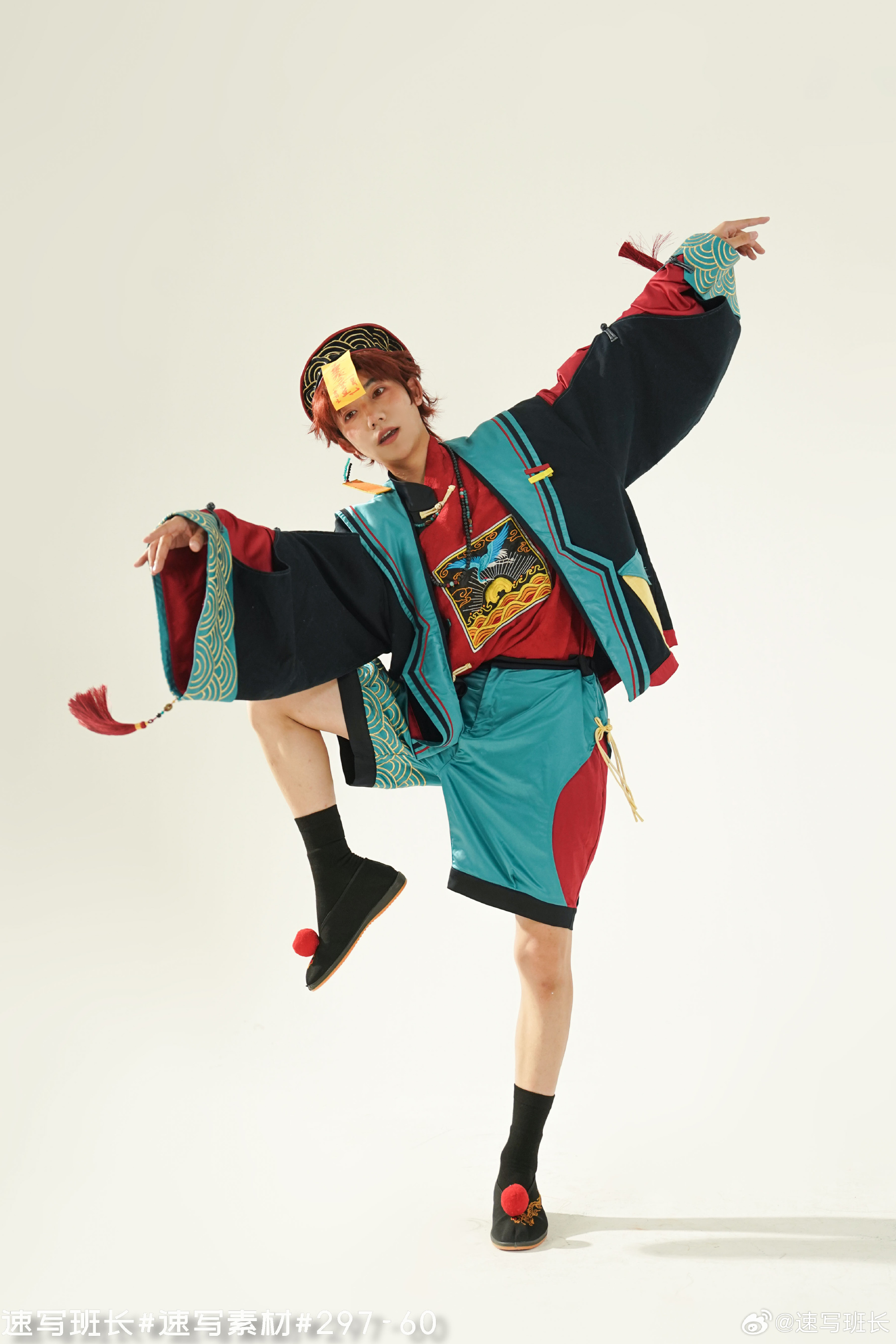 a young man with reddish brown hair is striking a dynamic dance pose. he is wearing a vibrant, eclectic outfit that blends traditional asian elements with modern streetwear. the top is a black jacket with wide sleeves, decorated with a gold and red pattern. underneath, he wears a red tshirt with a colorful graphic. his shorts are teal with red accents. he has black socks and slipon shoes with red pompoms. the background is a clean white, suggesting a studio environment. the lighting is bright and even, highlighting the details of the clothing and the pose.