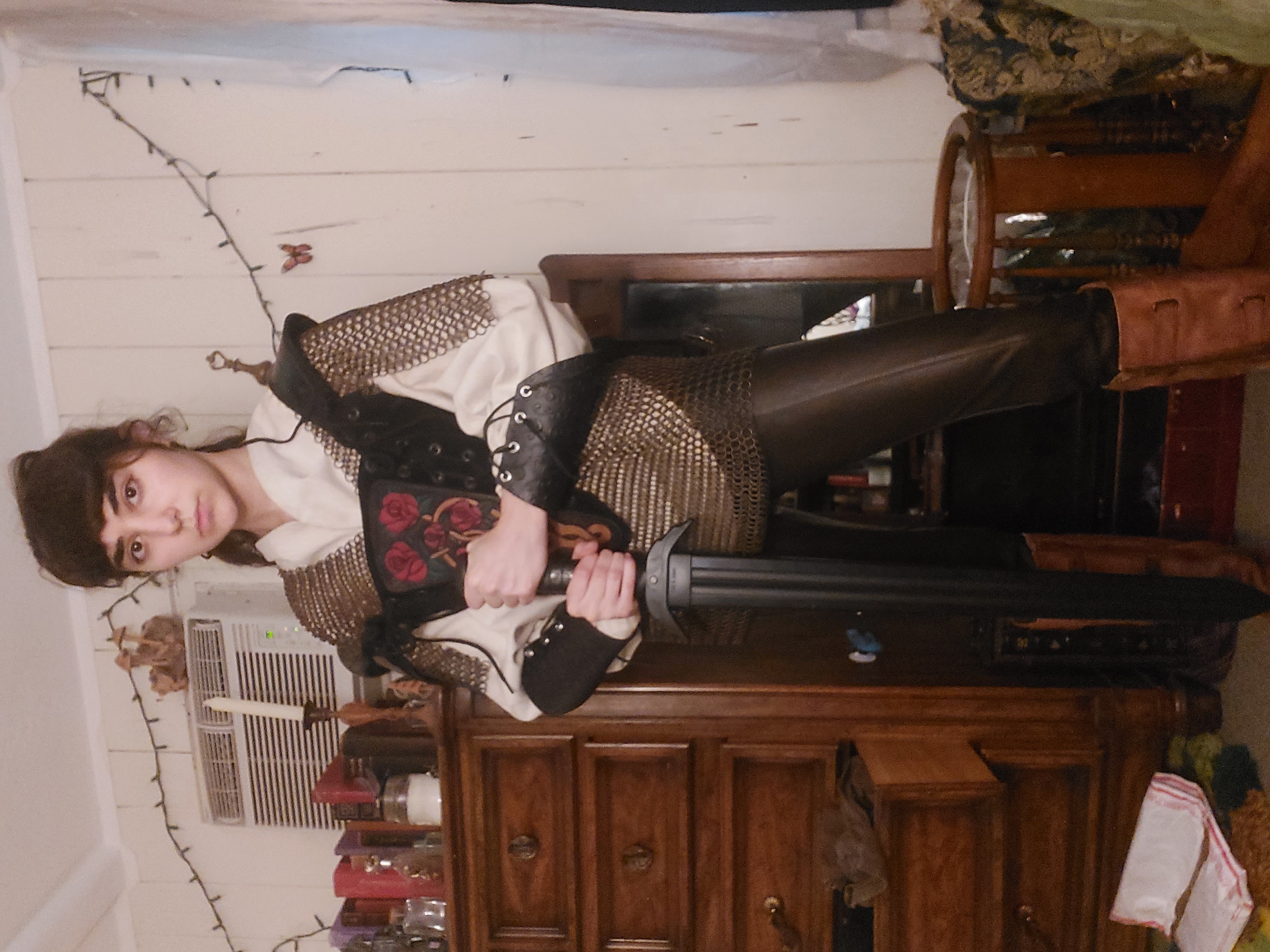 a young woman, possibly a teenager, is dressed in a medievalinspired fantasy costume. she's wearing a creamcolored shirt over chainmail, dark leather pants, and tall boots. the chainmail has an embroidered rose design. she's holding a sword and standing in a room with antiquestyle wooden furniture. the overall ambiance is dark and mysterious, suggesting a medieval or fantasy setting. the costume is detailed, showing attention to historical accuracy. the girl's serious expression enhances the roleplaying aspects of the scene.