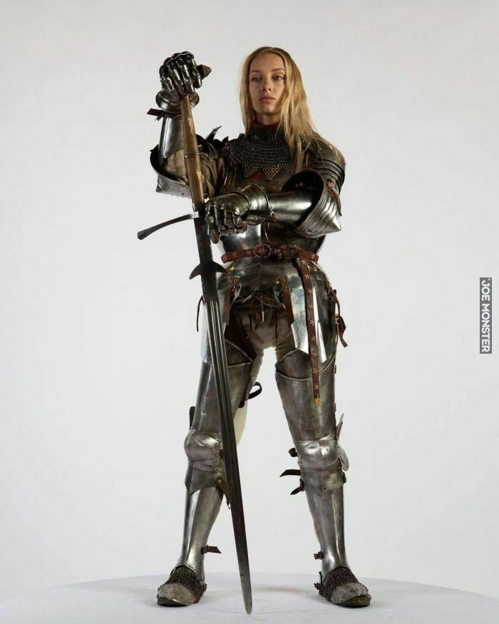 the image shows a female model in full medieval plate armor, standing against a plain white background. she holds a longsword upright, her grip firm, and maintains a confident pose. the armor is meticulously detailed, showing the intricacy of the metalwork and the chainmail underneath. the model's blonde hair contrasts against the metallic tones of the armor.  the overall impression is one of strength, power, and historical accuracy.  the image appears to be a professional studio shot, possibly for historical reenactment or a similar purpose.