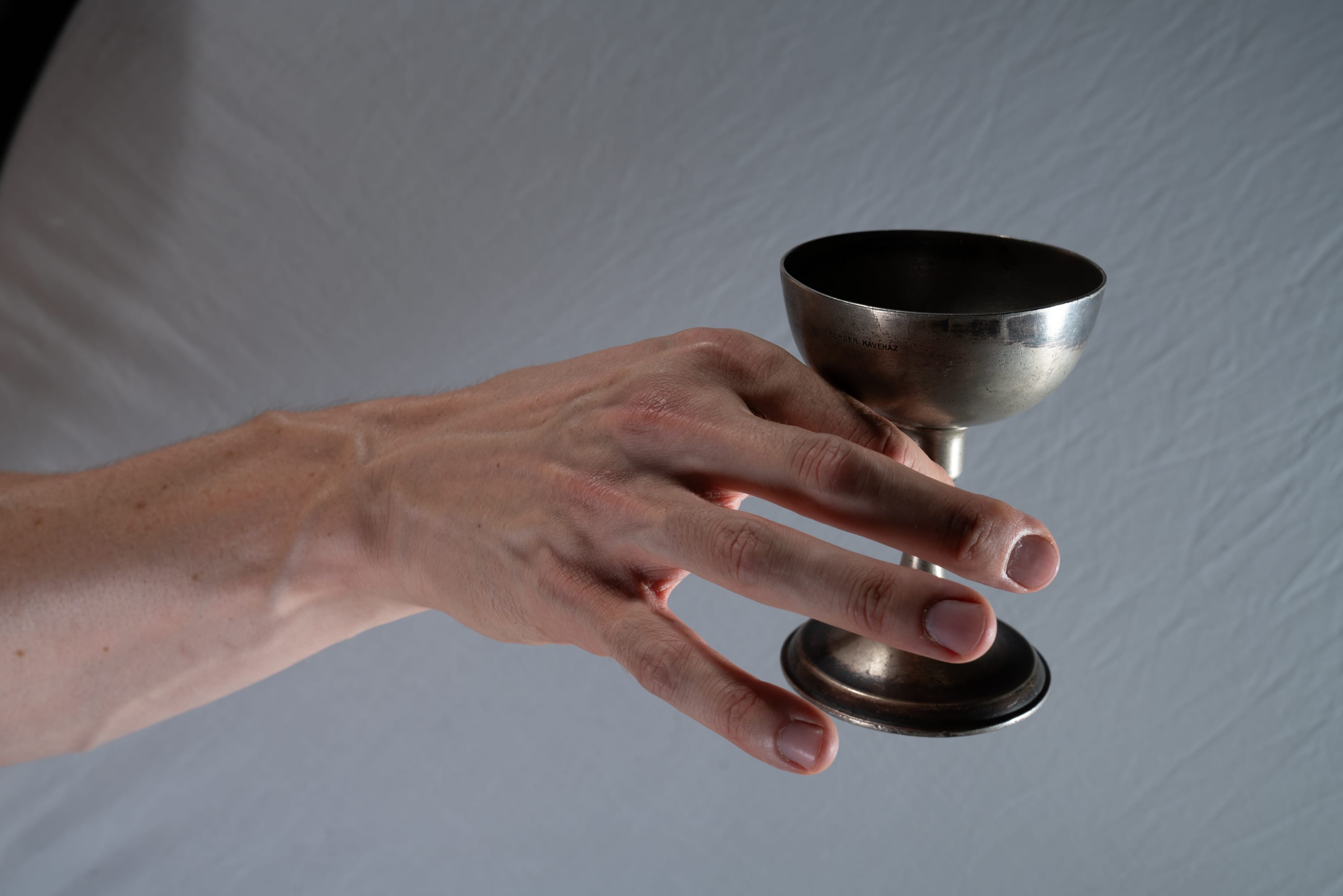 a closeup image featuring a fairskinned hand elegantly holding a gleaming silver chalice against a subtly textured gray backdrop. the chalice, crafted from polished silver, exudes an air of antiquity and ceremonial significance. the soft lighting accentuates the smooth curves of the chalice and the delicate details of the hand, highlighting its veins and contours.