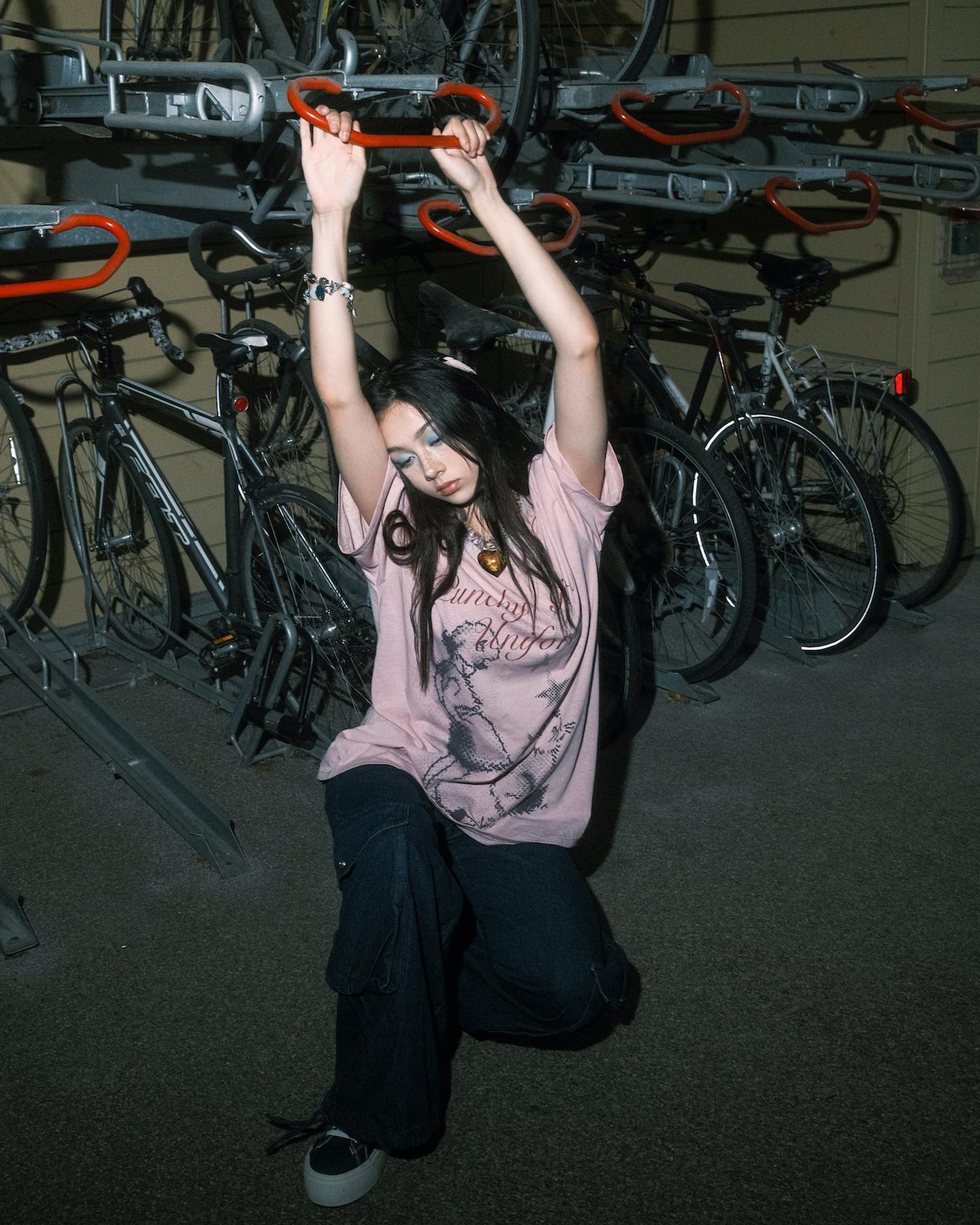 a young asian woman, possibly a teenager, is captured in a moody, candid portrait. she is posing leaning against a metal bike rack filled with parked bicycles in an urban setting at night. her clothing style is grungeinspired, with a pink shirt and dark cargo pants. the overall atmosphere is dark and cool, enhancing the alternative fashion and the model's attitude. the image has a street style feel, and the contrast between the dark environment and the girl's light shirt makes her stand out.