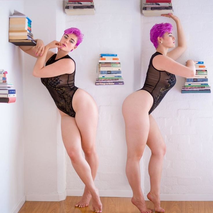 the image shows two fullbody shots of a woman with pink hair wearing a black bodysuit. she is posing indoors against a white brick wall with shelves of books. the woman is barefoot in both shots, with a hardwood floor beneath her. she has a curvaceous figure and is presented in a body positive way. the background features stacks of books of varying sizes and colors on small shelves attached to the wall.