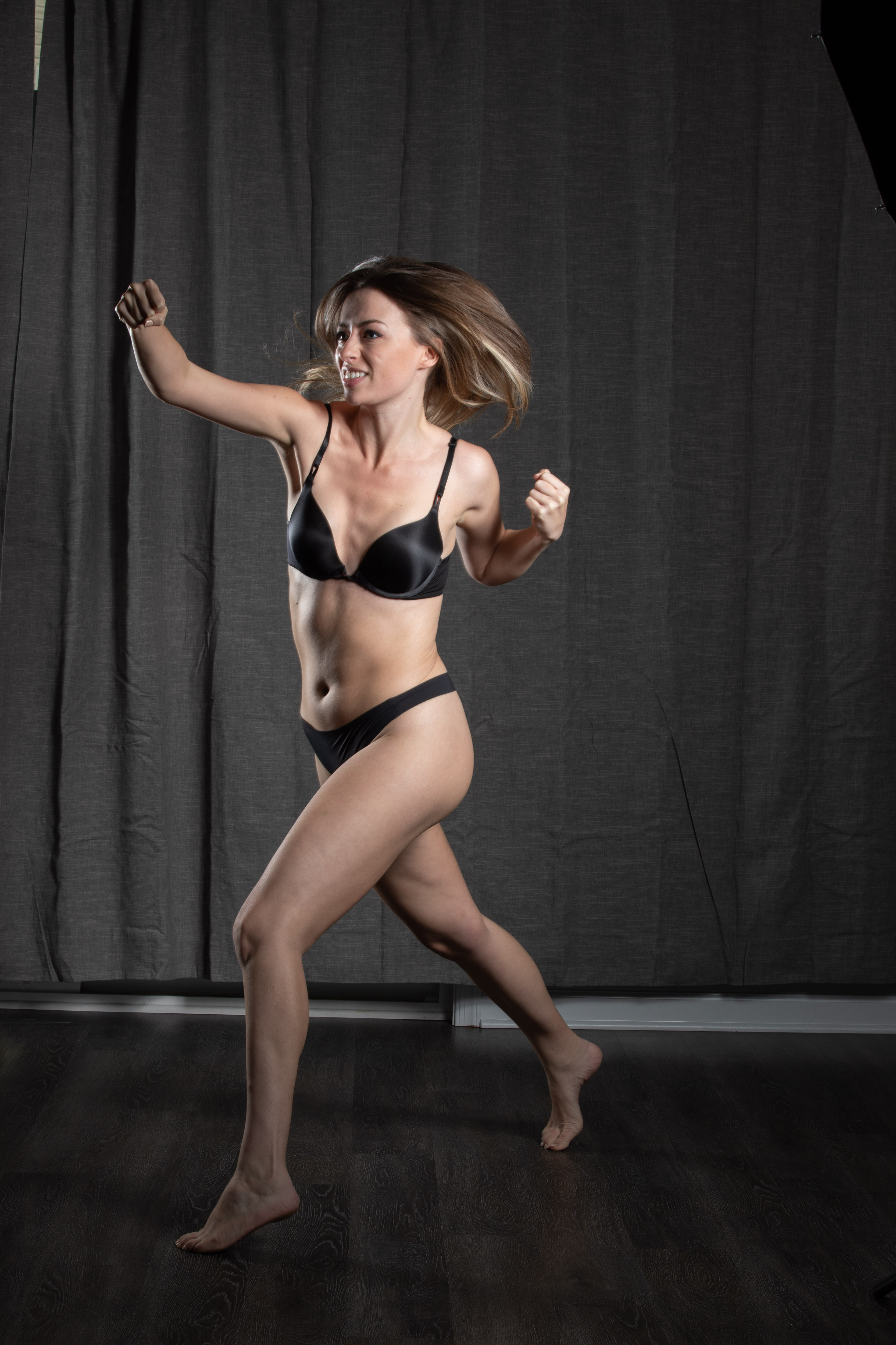 a woman with long brown hair is captured midrun against a dark gray backdrop. she is wearing black underwear and has a determined expression on her face. her arms are raised as if she is pumping them while running, and her hair is flowing behind her. the image conveys a sense of energy and athleticism. the lighting is artificial, likely from a studio setup, and illuminates her figure against the dark background.