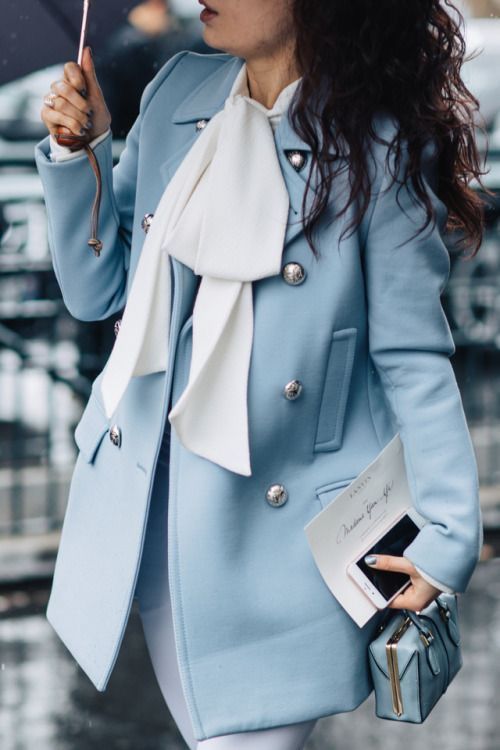 a stylish woman is seen outdoors on a rainy day, holding an umbrella and showcasing a sophisticated look. she wears a light blue doublebreasted coat with silver buttons, a crisp white blouse with a bow tie, and white pants. her accessories include a small light blue box bag and a smartphone. the overall style is elegant and classy.