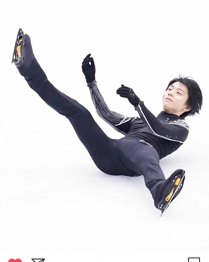 a figure skater in black pants and a black and white jacket falls on the ice while attempting a jump during a competition. his right leg is stretched out behind him, while his left leg is bent at the knee and he is lying on his stomach with his arms outstretched. the ice surface is white and the skater is wearing a black helmet.