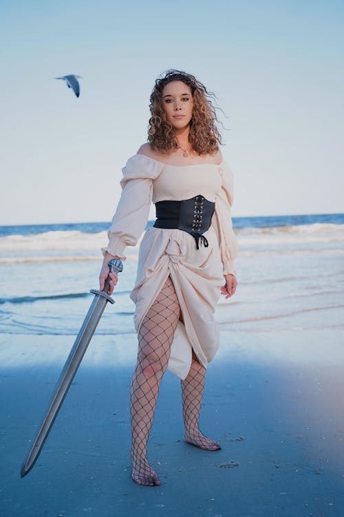 a full shot of a young woman dressed in a creamcolored offtheshoulder dress and black corset, holding a large sword. she is standing on a sandy beach, near the ocean waves. she is wearing fishnet stockings and is barefoot. a seagull is visible in the upper left corner of the image. the sky is clear and blue. the woman is looking directly at the camera with a serious expression. the overall mood of the image is dramatic and slightly mysterious.