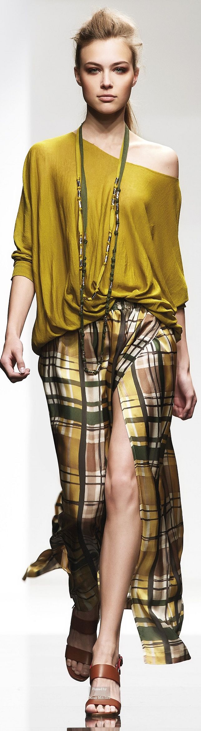 a woman in a yellow top with a plaid skirt is posing on a runway during a fashion show. she is wearing a gold necklace and brown sandals. she has her hair styled up and her eyes are looking at the camera.