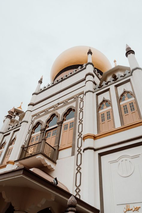 a lowangle, closeup shot of the masjid sultan in singapore. the image highlights the mosque's intricate details, including its golden dome, white walls, brown wooden doors and windows, and ornate decorative elements. the photograph captures the architectural beauty and grandeur of the islamic religious building. the sky is overcast.
