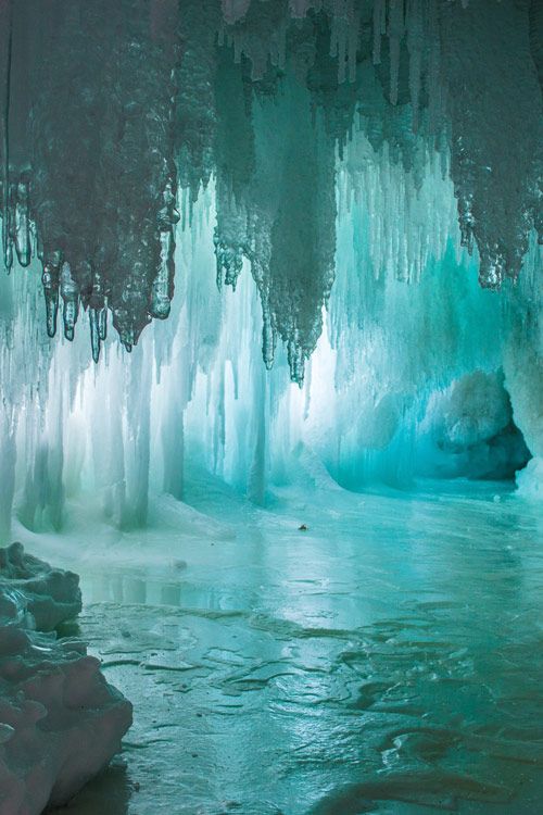 this is a photograph of an ice cave with numerous icicles hanging from the ceiling. the cave is filled with ice formations and some open water, creating a cold and otherworldly landscape. the predominant colors are blue and white, emphasizing the cold and icy nature of the environment.