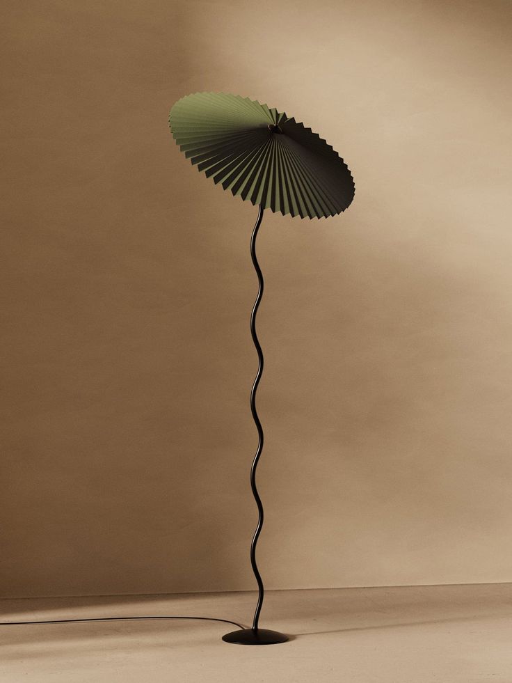 the image showcases a modern, minimalist floor lamp. the lamp features a unique pleated lampshade in a muted olive green color that resembles an umbrella in shape.  the shade is mounted on a long, slender, black stem that curves gently, adding to its elegant and somewhat whimsical design. the lamp sits on a small, circular black base.  the background is a neutral, light beige or tan, allowing the lamp's design and color to stand out. the overall aesthetic is clean, sophisticated, and calming.