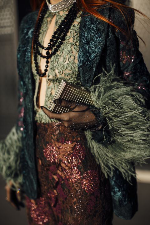 a closeup shot of a woman dressed in a luxurious, richly textured outfit.  she's wearing a dark teal velvet jacket adorned with feathers, a delicate green lace top, and a shimmering burgundy sequined skirt.  she accessorizes with a multistrand black beaded necklace, a sparkling rhinestone choker, black lace gloves, and a metallic gold rectangular clutch. the overall style evokes a sense of vintage glamour and sophistication. the focus is on the intricate details of the clothing and accessories, highlighting the rich fabrics and textures.  the woman's red hair peeks out from under the jacket, adding a vibrant contrast to the dark tones of her ensemble.