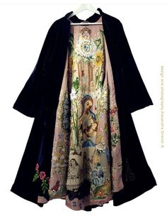 a black velvet cape and a long, multicolored floral print dress is draped over a hanger. the dress has a pink base with a variety of embroidery that includes images of birds, angels and flowers. the black velvet cape has a flowing, loose fit and adds a touch of drama and mystery to the already unique ensemble.