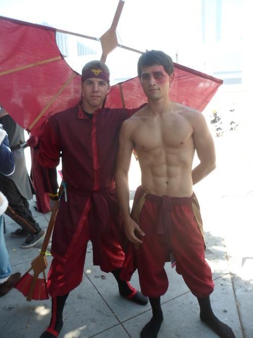 two male cosplayers dressed as characters from the cartoon show avatar the last airbender, aang and zuko. aang is on the left holding a fire nation fan and zuko is on the right holding a sword.  the cosplayers are posed for a photograph.  they are both wearing their iconic red and orange attire with a red and orange backdrop.  they are standing in front of a building in the background.