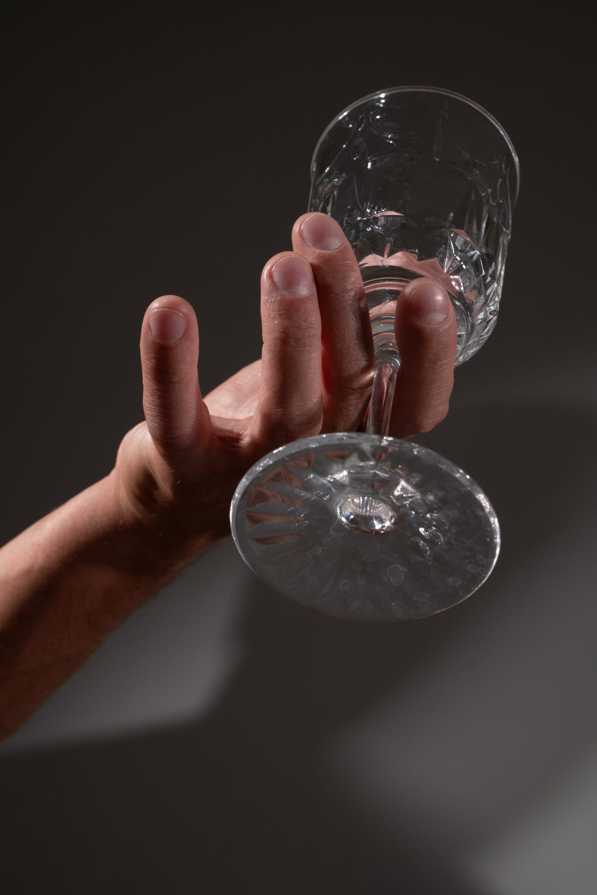 a closeup shot of a hand holding up a clear crystal wine glass against a dark gray background. the glass is empty and the bottom of the glass is facing the camera. the hand is holding the stem of the glass with four fingers around the stem and the thumb resting on the side of the glass. the lighting is soft and diffused.
