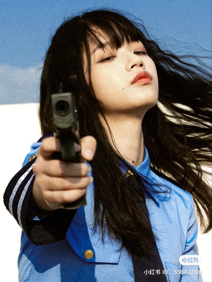 a woman with dark hair and bangs points a gun at the viewer. she is wearing a blue shirt with a tie, possibly a police uniform. the background is a blurry white and blue.