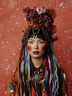 a woman with long colorful hair and a flower crown on her head.  she is wearing colorful clothing and a necklace with a large pendant. she has a colorful makeup, and her eyes are looking at the camera. the background is red and blurred.