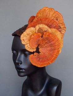 a woman's mannequin with a striking orange mushroom as a hair accessory.  the mushroom is orange and red and looks like a hairpiece.  it covers the top of the head and curls around the side, creating a unique and bold statement.  the mannequin is also wearing a black dress that is simple and does not detract from the mushroom headpiece. the backdrop is light grey. the overall image is creative and artistic.